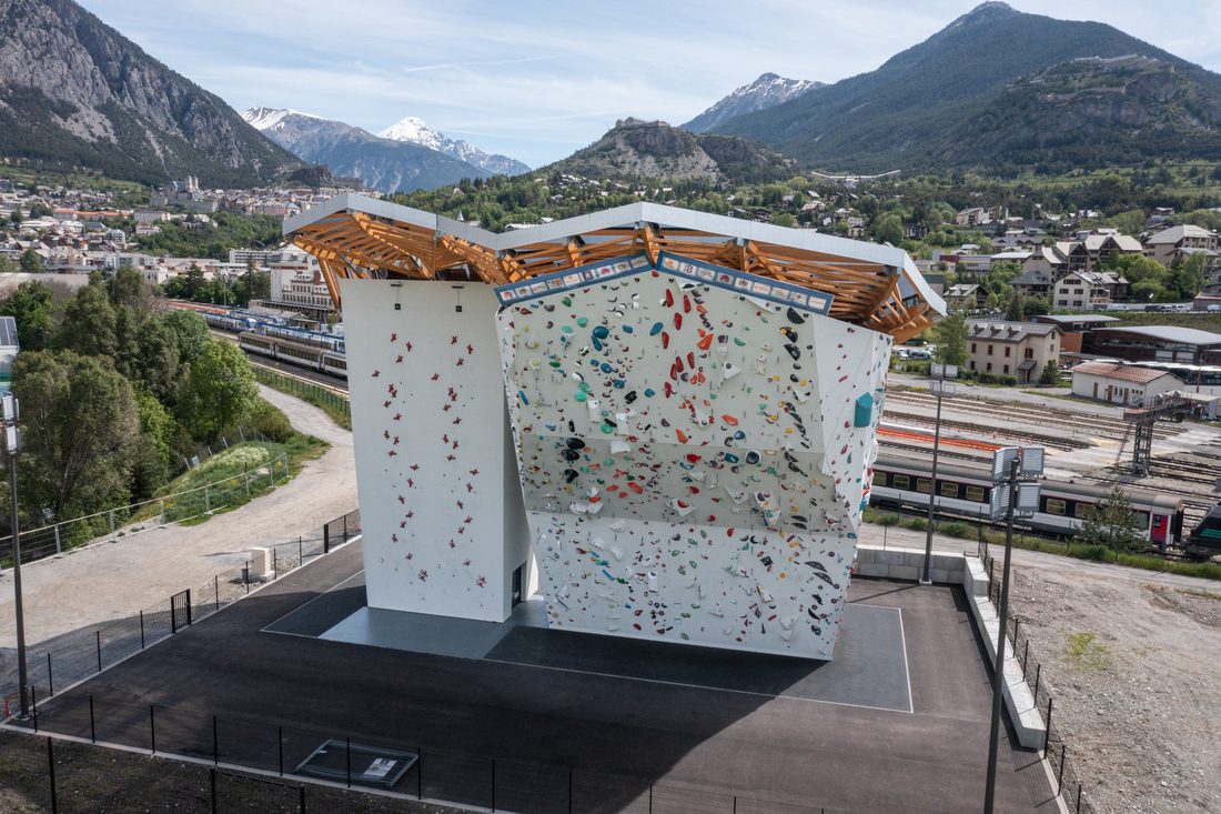 Outdoor climbing wall with a transparent canopy set in a mountain landscape, combining architecture and nature.