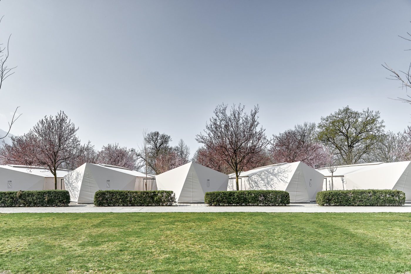 Modular textile shelters integrated into a landscaped park with blooming trees.