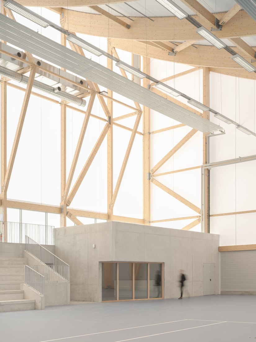 Détail intérieur d’un gymnase avec ossature bois, murs en membrane textile translucide et équipements techniques apparents.