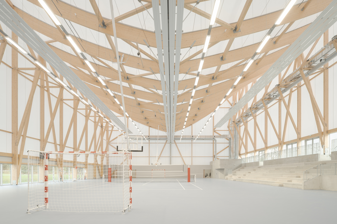 Intérieur d’un gymnase avec charpente en bois apparente et façade textile translucide laissant entrer la lumière naturelle, réalisée avec la membrane Tenseo 1002 S2.