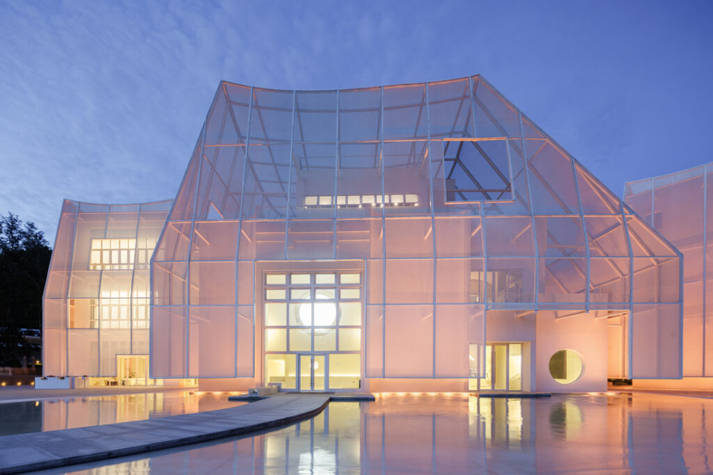 Façade en membrane textile translucide enveloppant un bâtiment aux volumes géométriques, illuminé au crépuscule et se reflétant dans un bassin.