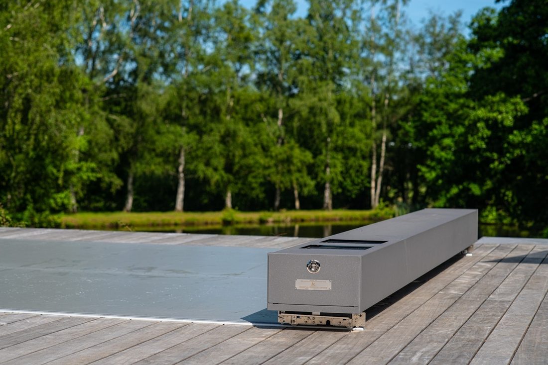 Coffre motorisé gris installé en bout de piscine sur terrasse en bois, avec plan d’eau et arbres en arrière-plan.