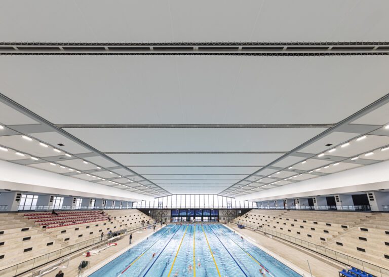 Bassin olympique intérieur avec plafond textile tendu et gradins en pierre.