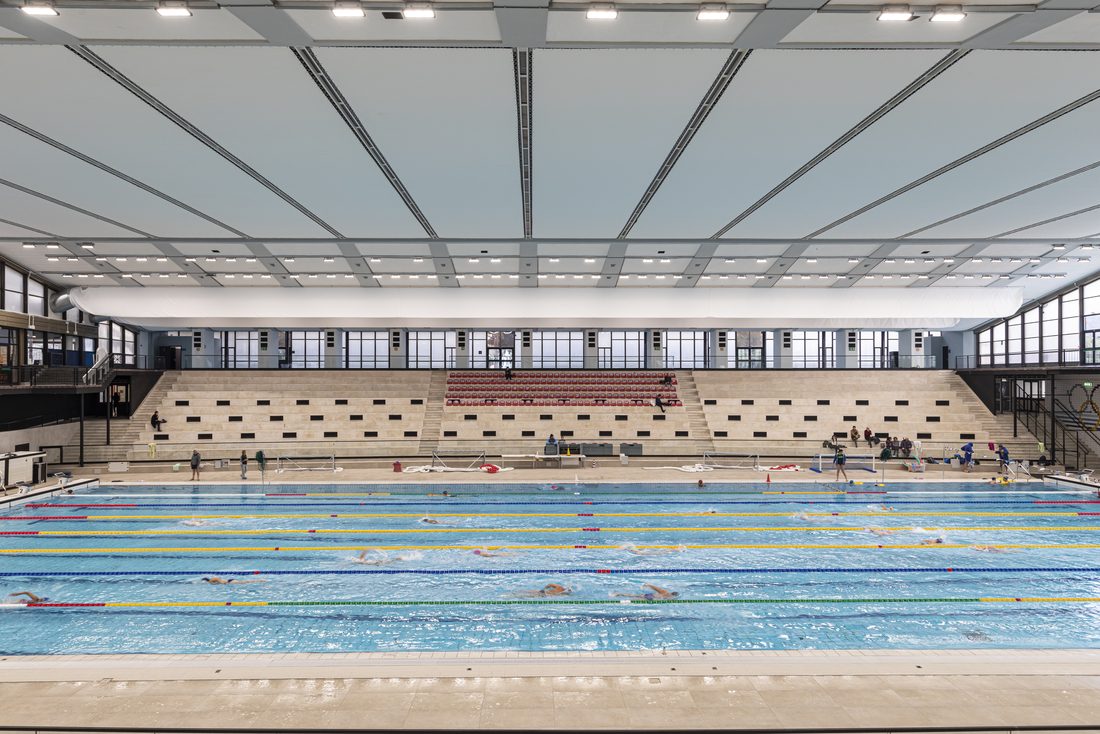 Piscine olympique intérieure avec plafond acoustique en membrane textile Serge Ferrari, éclairage intégré et tribunes.