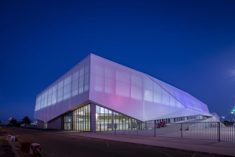 Façade de stade en membrane composite translucide, rétroéclairée, formant un volume architectural lumineux de nuit.