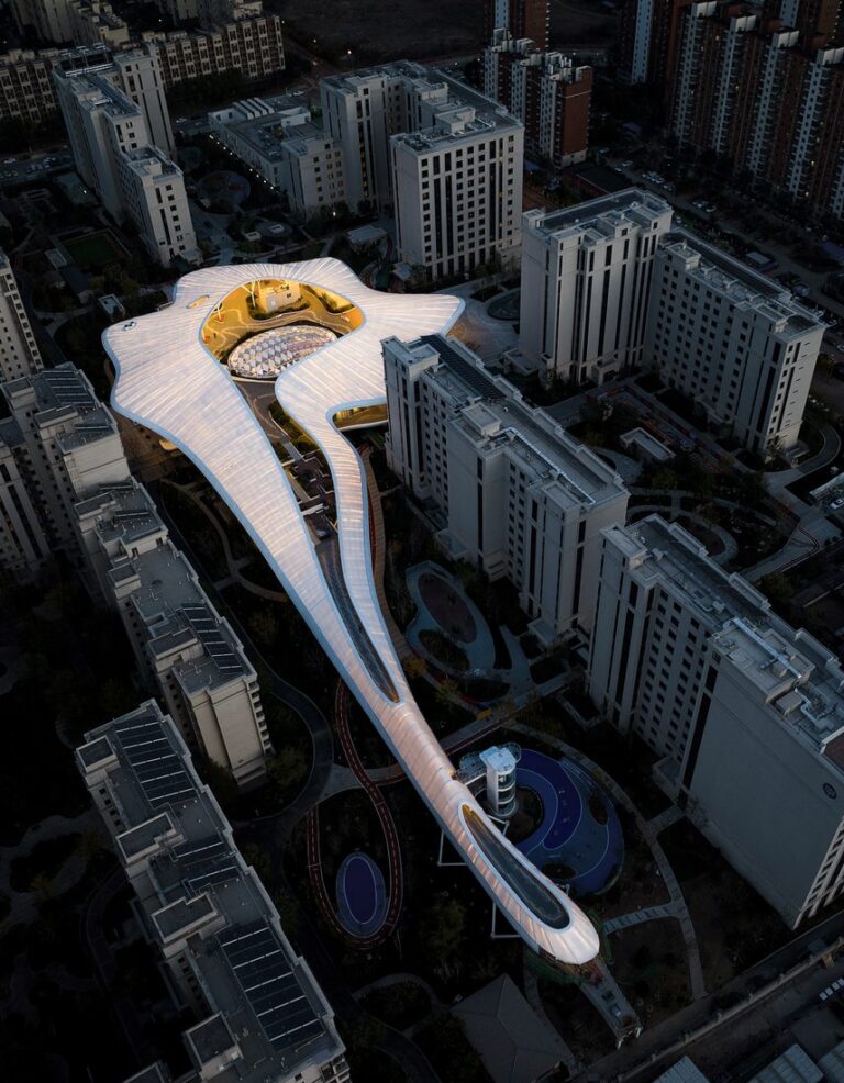 Vue aérienne de nuit d’un bâtiment contemporain en forme organique, éclairé, au milieu d’immeubles résidentiels.