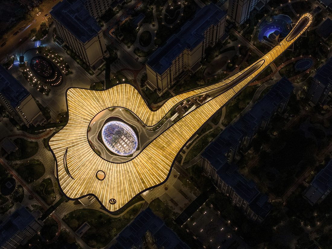 Vue aérienne au coucher du soleil d’un centre de soin au toit ondulant, entouré d’immeubles, avec un lac et des montagnes en arrière-plan.