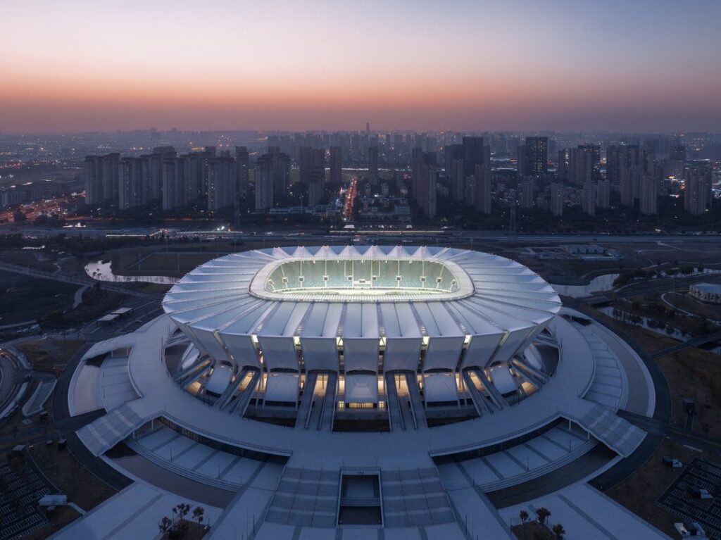 Vue aérienne au crépuscule d’un grand stade, dont la toiture annulaire en membrane composite encadre l’ouverture centrale au-dessus des tribunes, avec la skyline de la ville en arrière-plan.