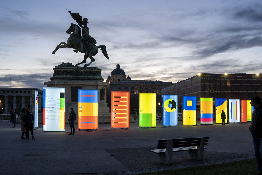 Exposition artistique en plein air avec toiles imprimées rétroéclairées, installée sur une place urbaine au crépuscule.