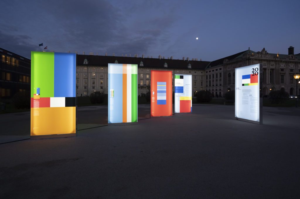 Installation artistique extérieure avec panneaux lumineux habillés de toiles imprimées colorées, sur une place au coucher du soleil.