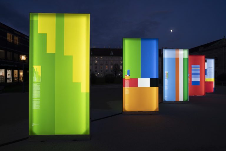 Exposition artistique en extérieur avec structures rétroéclairées recouvertes de toiles imprimées graphiques.