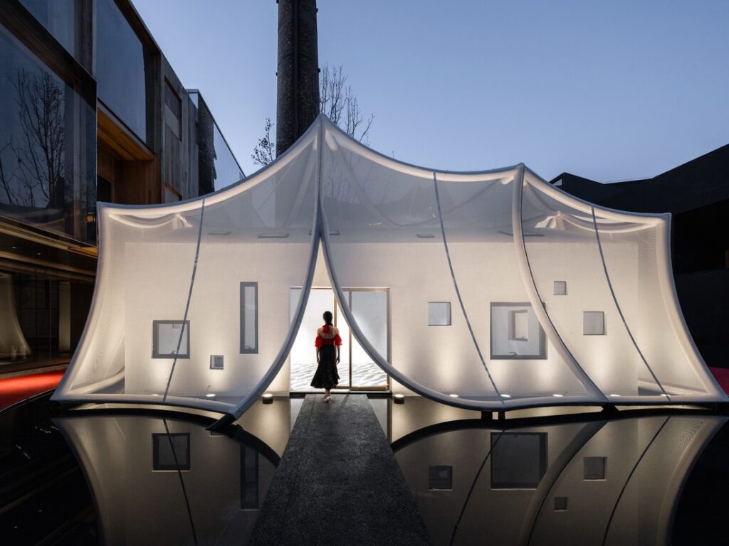 Pavillon contemporain recouvert d’une toile membrane translucide, éclairé de l’intérieur avec une silhouette entrant dans le bâtiment.