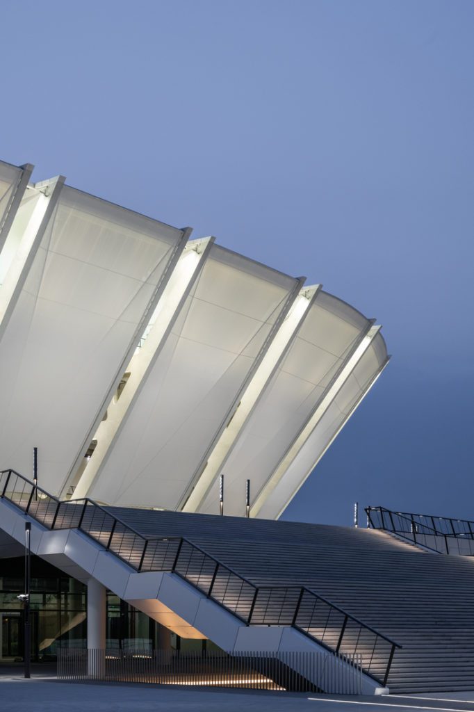 Vue en contre-plongée d’une façade de stade composée de panneaux de membrane blanche tendus entre nervures structurelles, au-dessus d’un large escalier extérieur.
