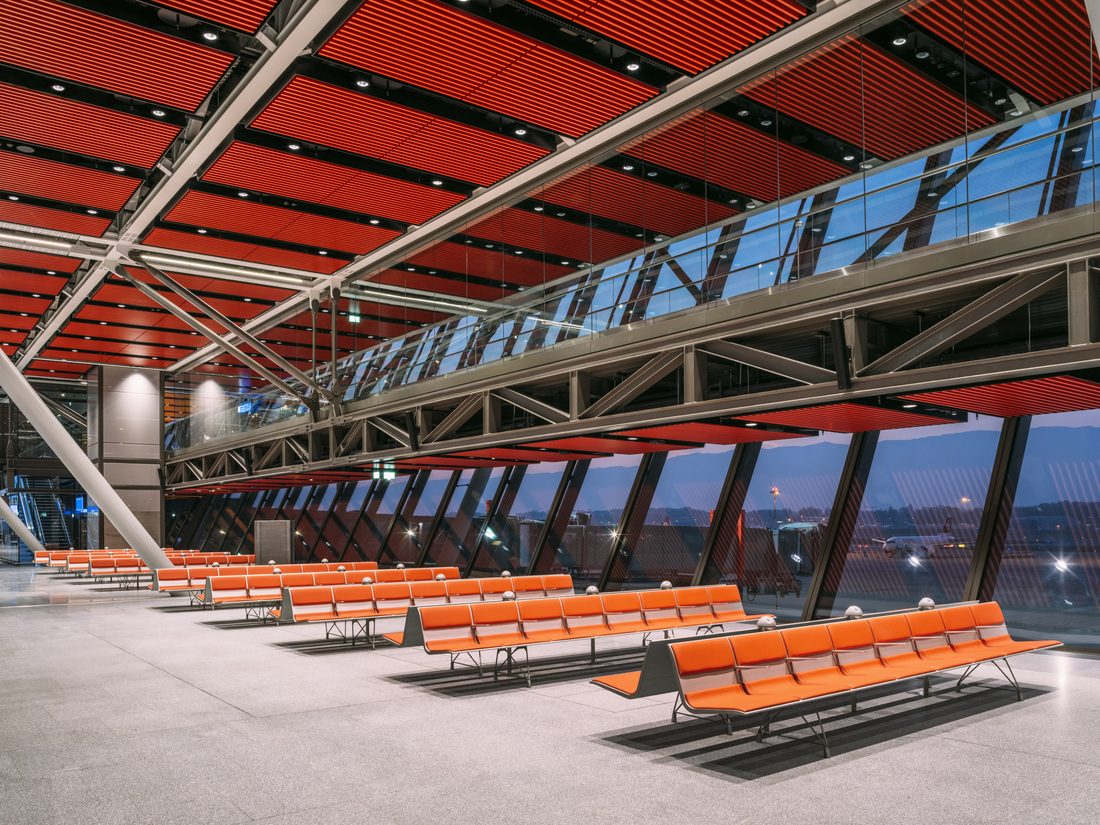 Rangées d’assises orange dans un terminal d’aéroport moderne avec structure métallique et plafond architectural rouge.