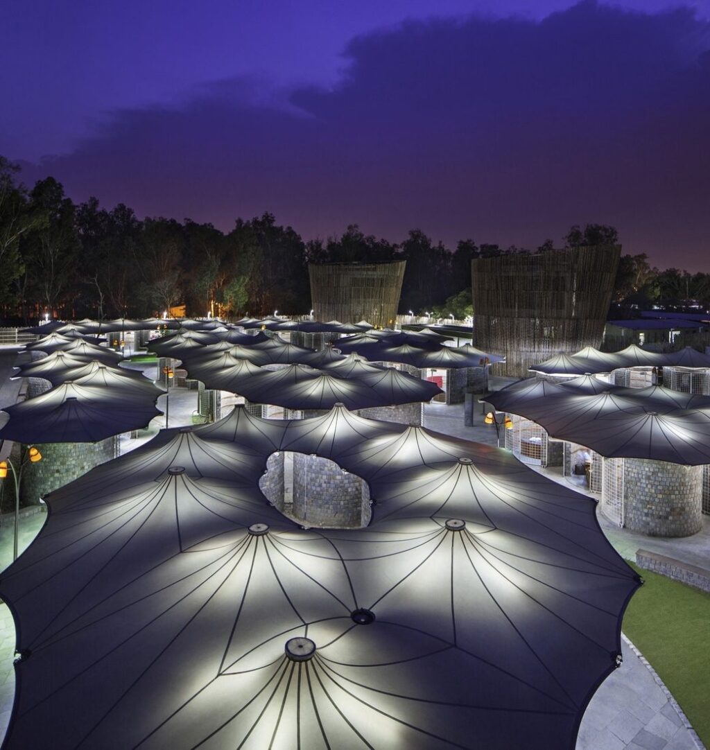 Vue de nuit d’un site public couvert par une multitude d’ombrières textiles en forme de parapluie, éclairées par en dessous.