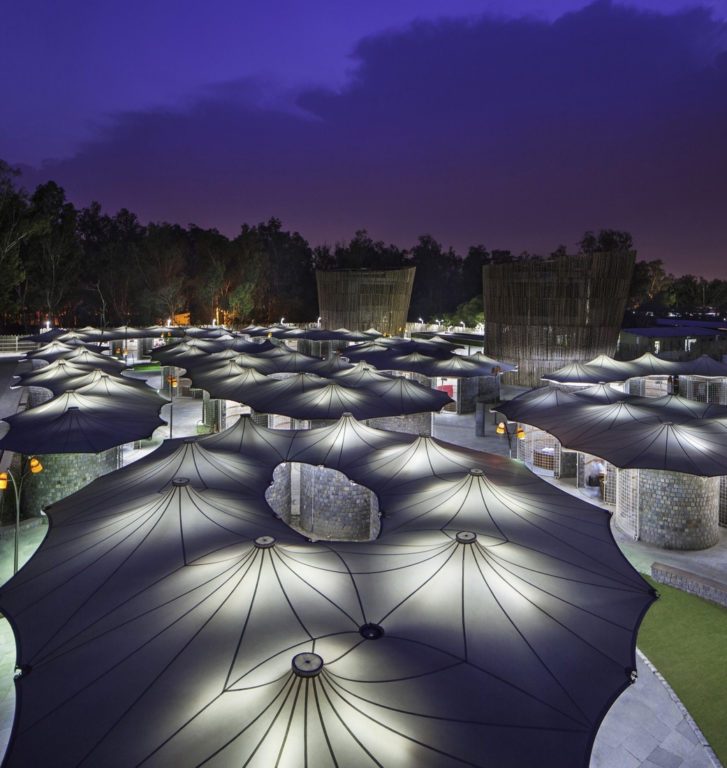 Vue de nuit d’un site public couvert par une multitude d’ombrières textiles en forme de parapluie, éclairées par en dessous.