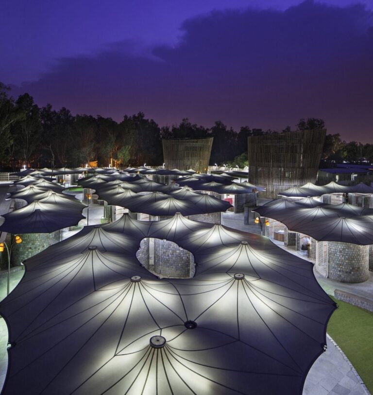 Vue de nuit d’un site public couvert par une multitude d’ombrières textiles en forme de parapluie, éclairées par en dessous.