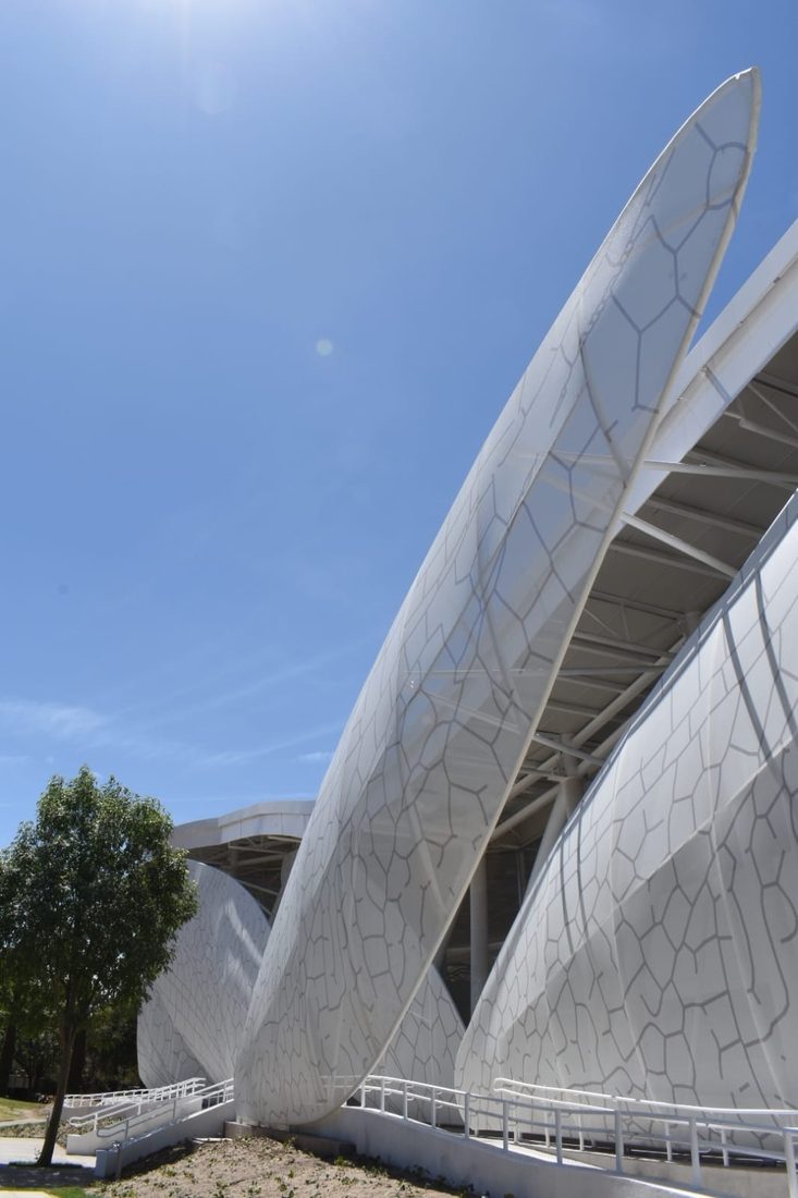 Close-up of a wing-shaped façade element clad with custom-printed Tenseo Frontside 381 textile mesh rising against a blue sky.