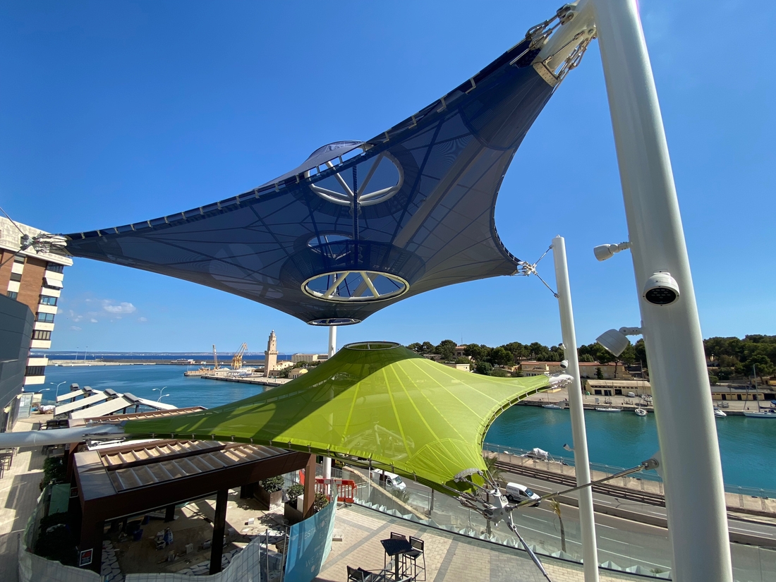 Voiles d'ombrage textiles surplombant les terrasses d'un centre commercial avec vue sur le port.