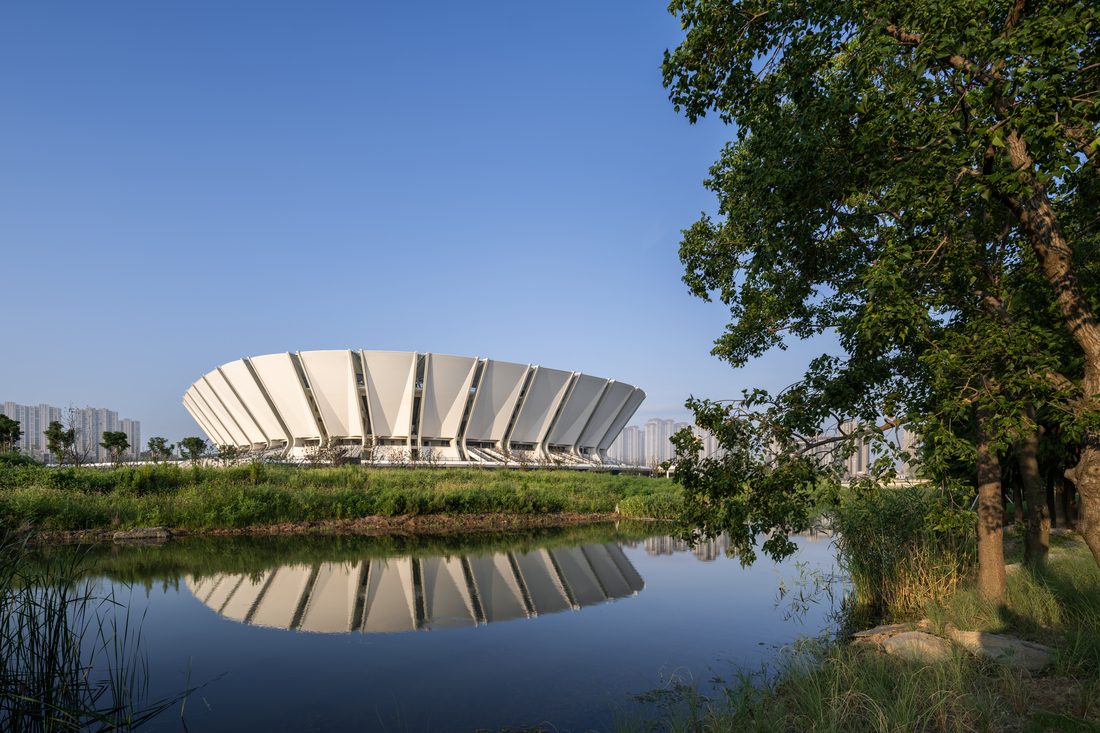 Side view of Kunshan Stadium reflected in a pond with trees in the foreground.