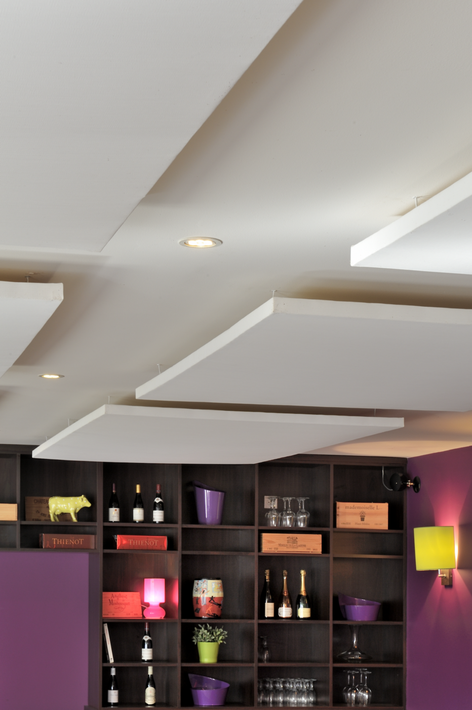 Plafond acoustique avec panneaux suspendus blancs dans un restaurant, au-dessus d’un mur d’étagères avec bouteilles et objets décoratifs.