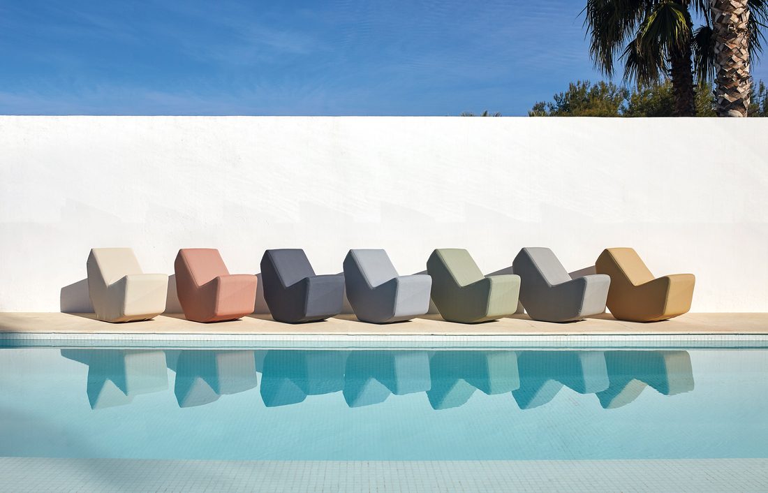 Alignement de fauteuils lounge colorés au bord d’une piscine avec mur blanc et ciel bleu.