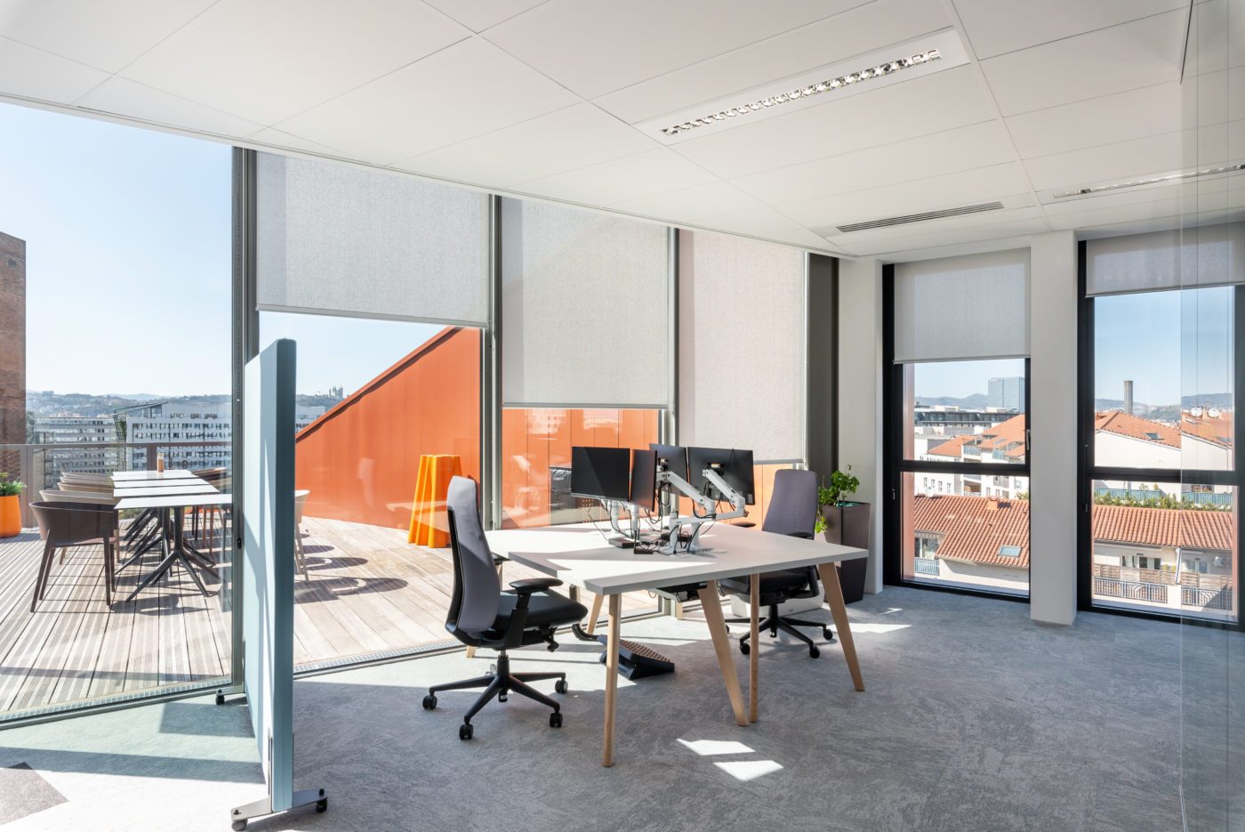 Bureaux lumineux équipés de stores enrouleurs intérieurs en toile technique Serge Ferrari sur grandes baies vitrées, avec vue sur une terrasse et la ville.