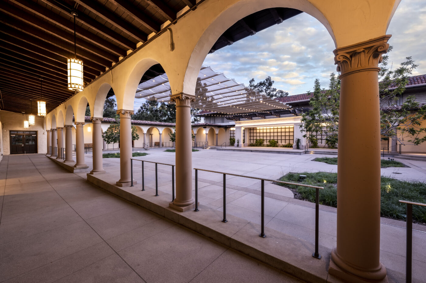 Arched colonnade with lit lanterns overlooking Cannon Plaza courtyard and a retractable shade canopy at dusk.