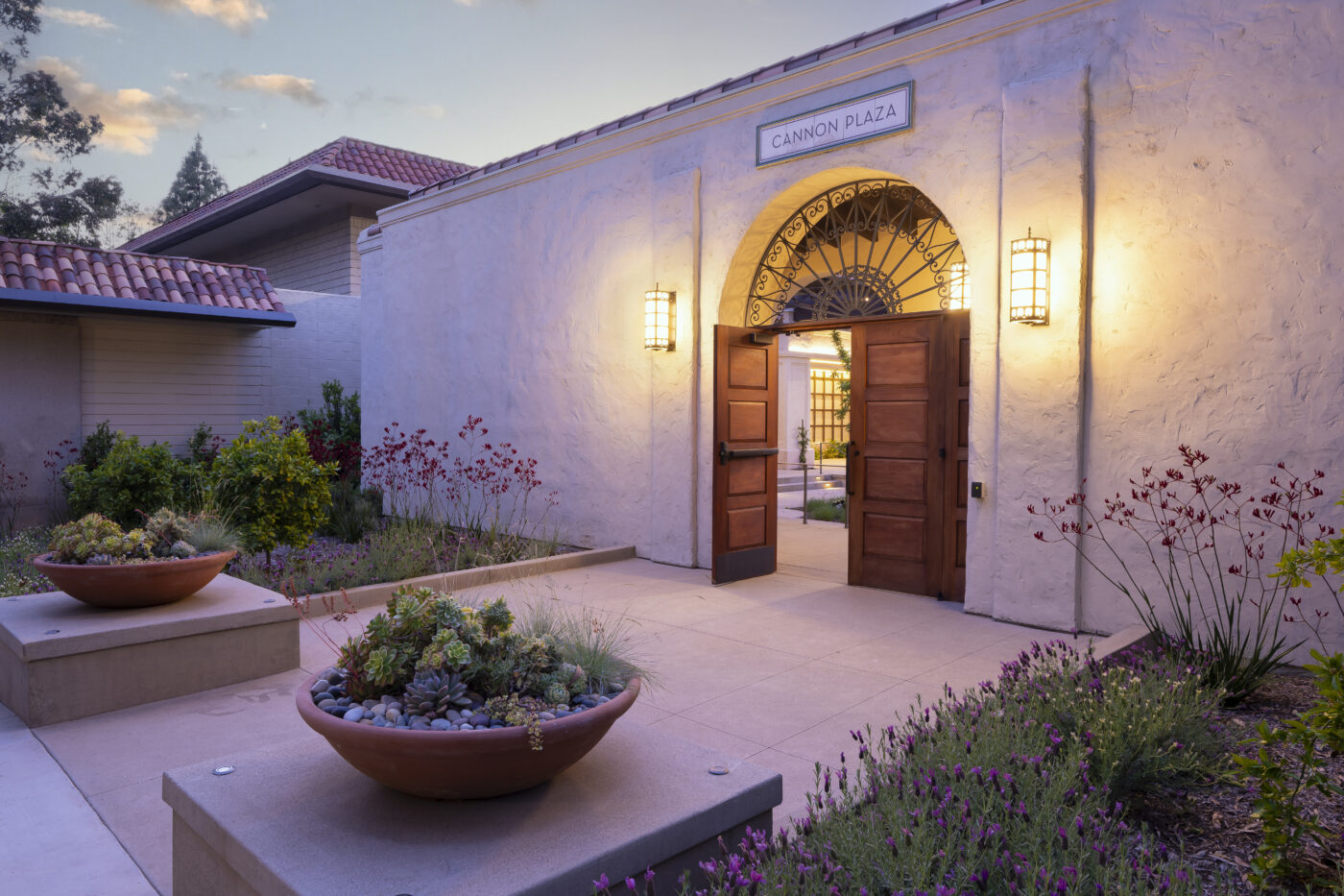 Open wooden doors beneath an arched entry labeled “CANNON PLAZA,” with wall lanterns and landscaped planters at dusk.