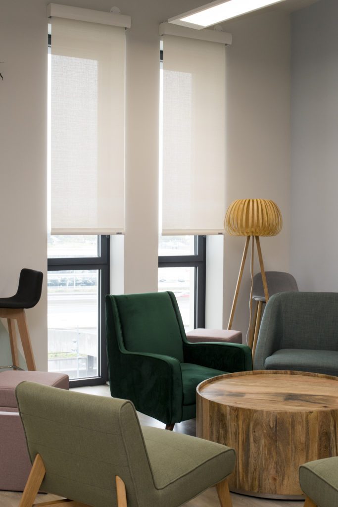 Espace lounge du projet INITIAL Griesser, avec fauteuils colorés, table basse en bois et stores enrouleurs blancs filtrant la lumière.
