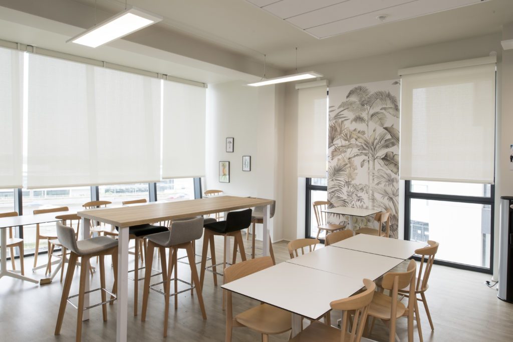 Espace de pause en entreprise avec grandes baies vitrées, tables hautes et basses, chaises en bois et stores enrouleurs blancs filtrant la lumière.