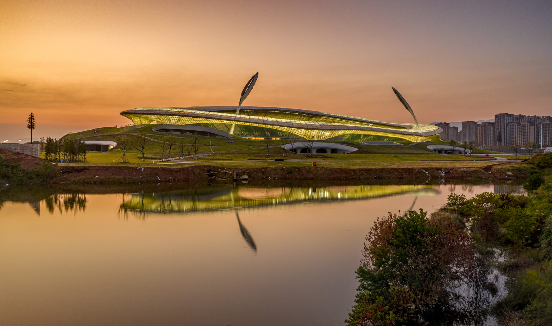 Quzhou Stadium au coucher du soleil avec façade lumineuse et toiture membrane Serge Ferrari reflétée dans l’eau