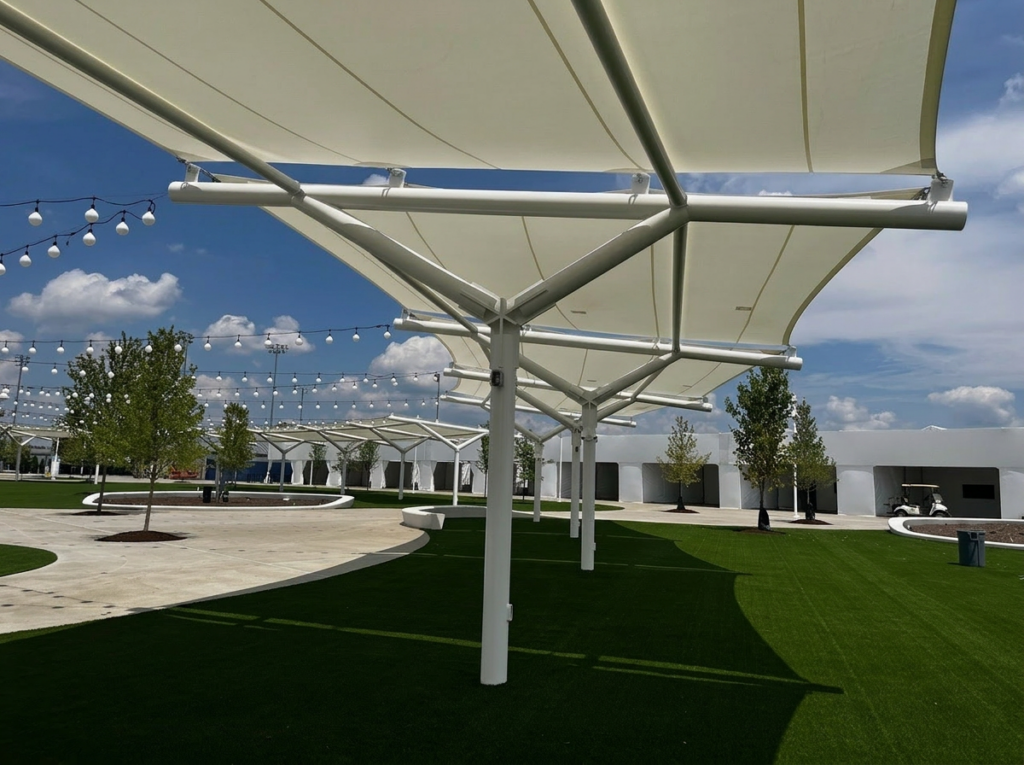 White shade sail structures over landscaped courtyard with pathways, grass, trees, and string lighting.