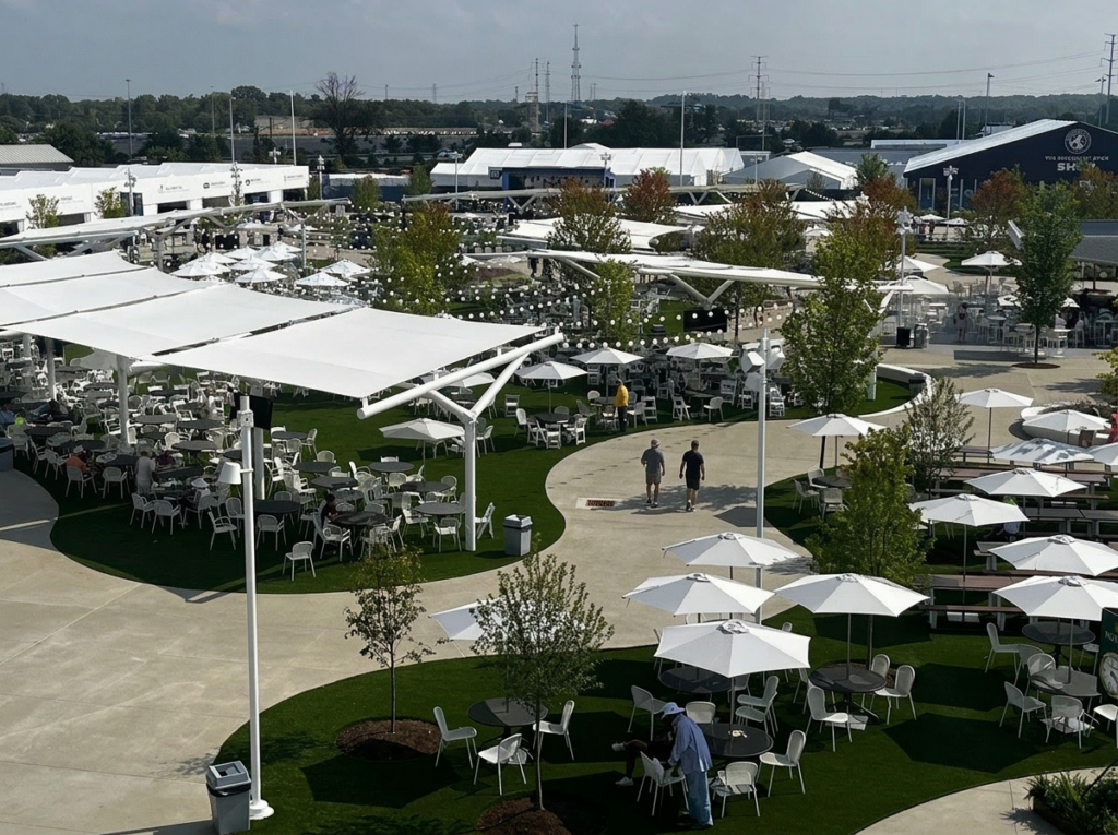 Large outdoor dining and event space with white shade sails, tables, umbrellas, landscaped paths, trees, and string lighting.