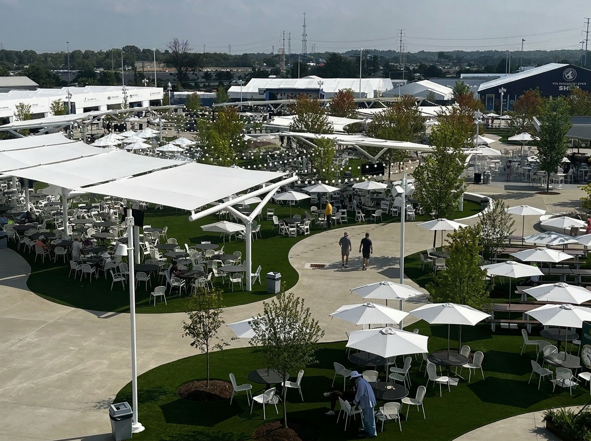 Large outdoor dining and event space with white shade sails, tables, umbrellas, landscaped paths, trees, and string lighting.