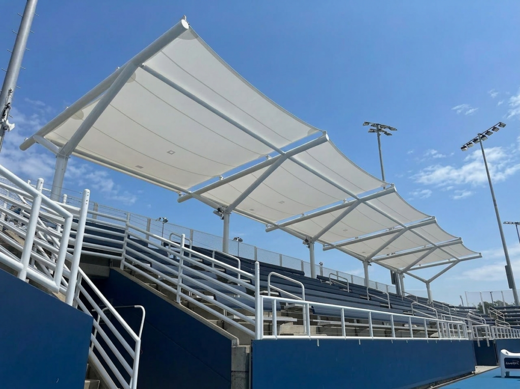 Tensile fabric canopy structure shading bleachers at an outdoor stadium with metal railings and lighting poles.