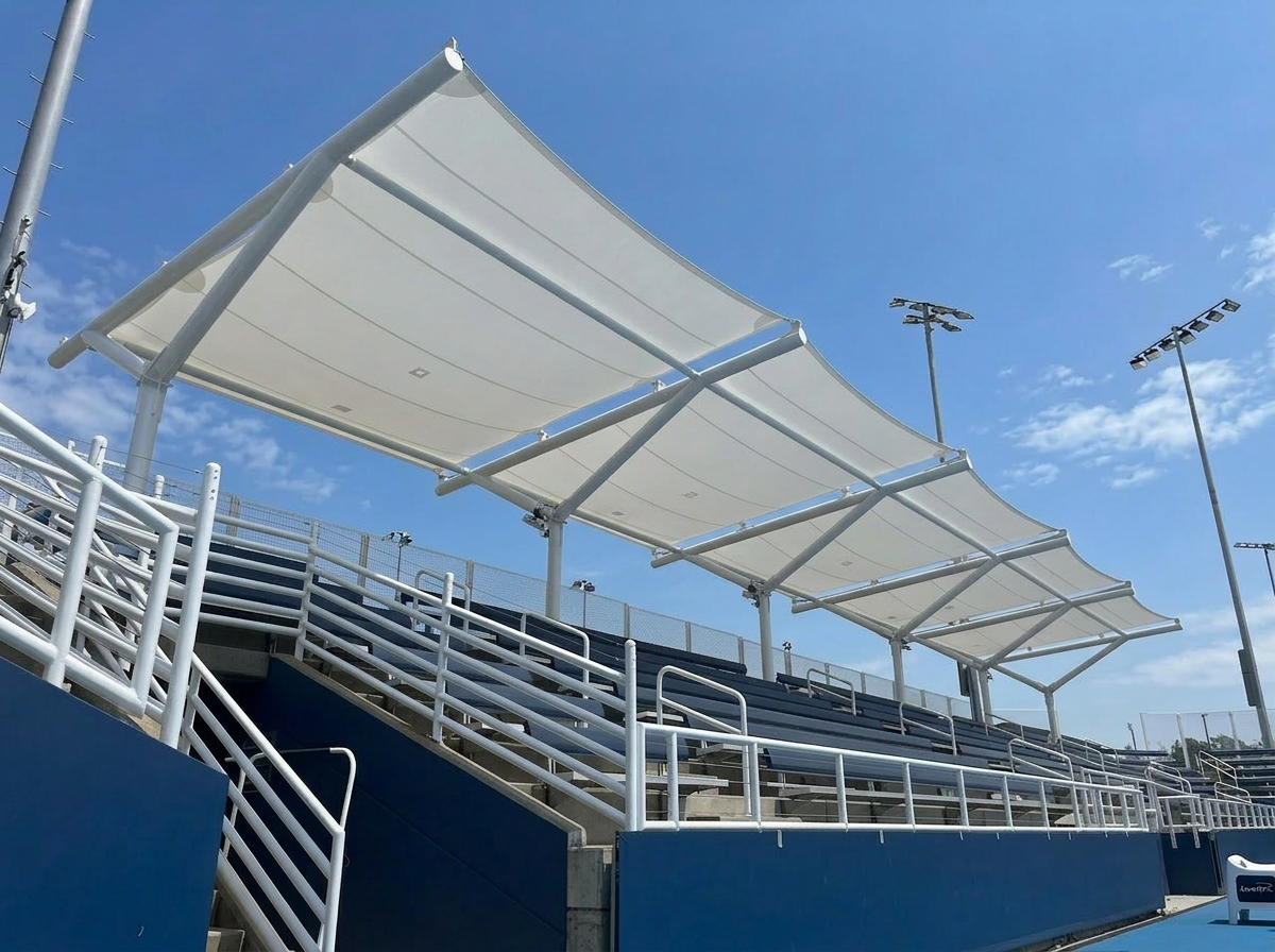 Tensile fabric canopy structure shading bleachers at an outdoor stadium with metal railings and lighting poles.