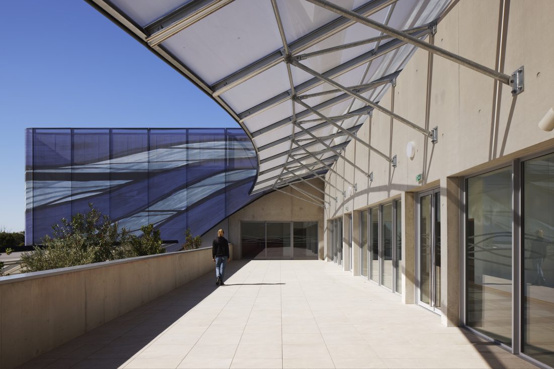 Terrace view of Sport Hall Nîmes with structural framing and printed mesh façade in the background.