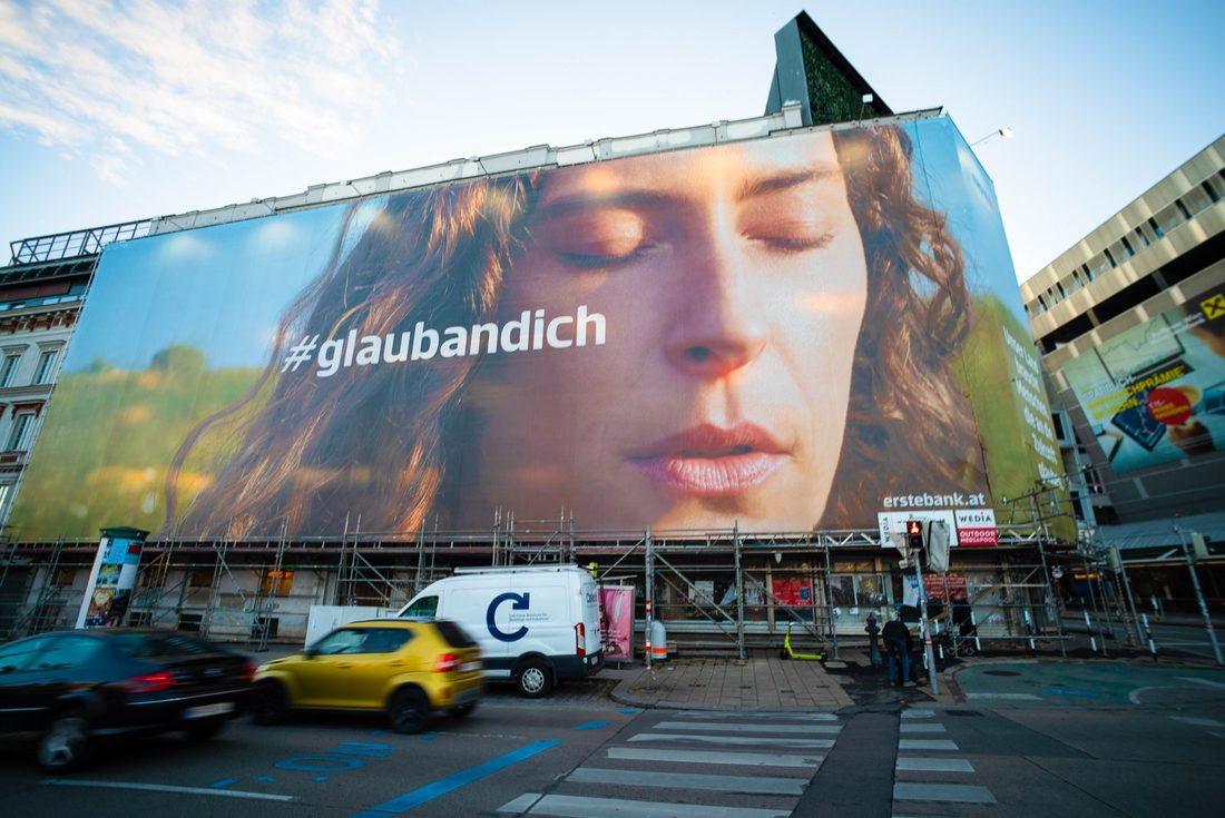 Bâche publicitaire grand format sur la façade d’un immeuble en ville, avec un portrait en gros plan et le message « #glaubandich », au-dessus d’une rue passante.