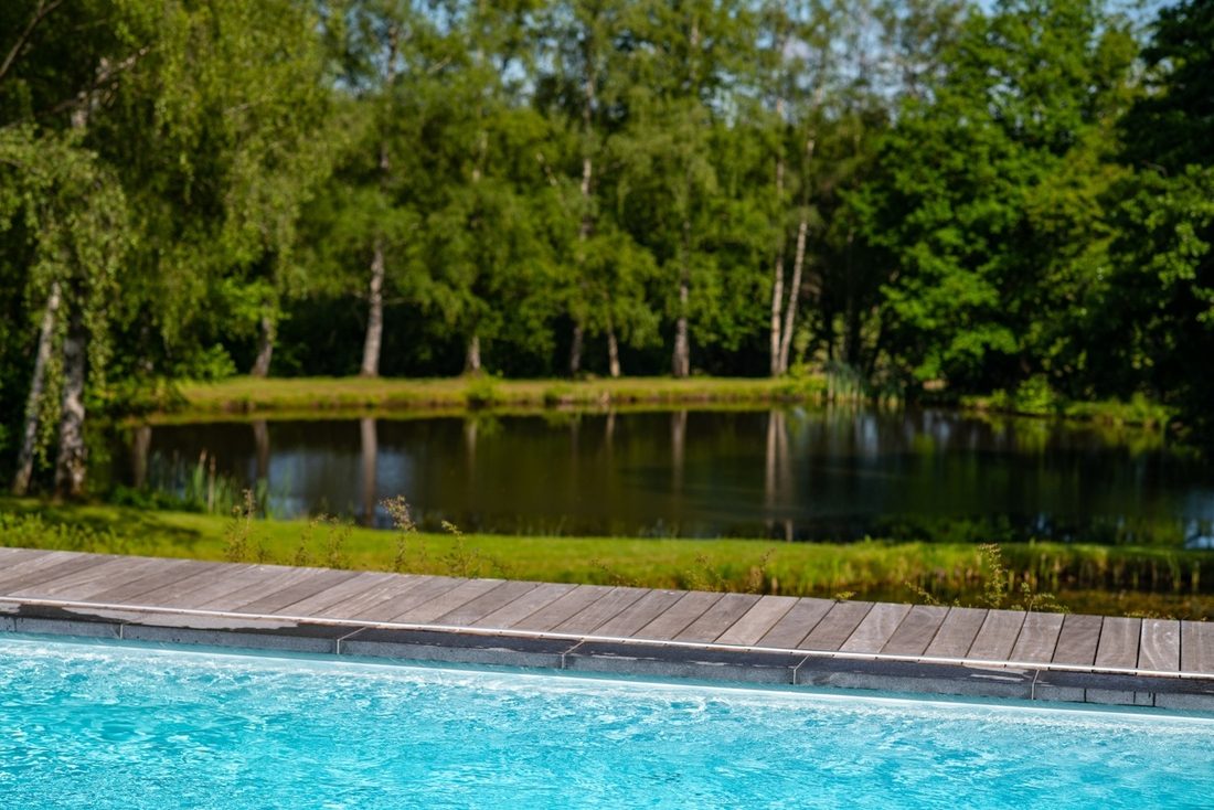Piscine extérieure avec eau turquoise bordée d’une terrasse bois, face à un étang et un paysage arboré.