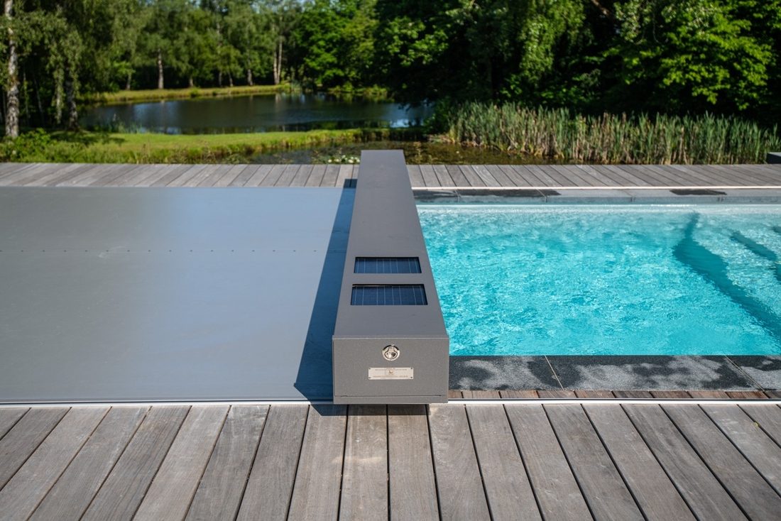 Piscine extérieure avec couverture automatique grise partiellement fermée, terrasse en bois et environnement arboré.