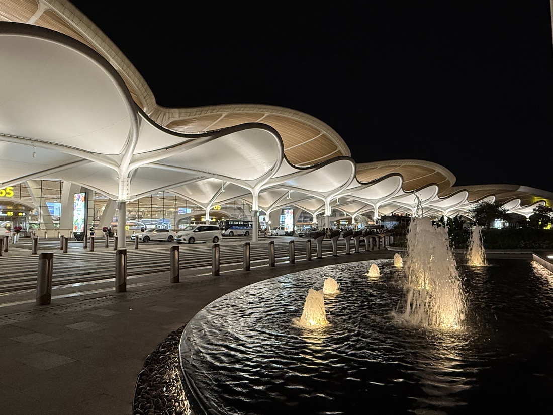 Membrane textile tendue éclairée de nuit, couvrant la zone de dépose devant un terminal, avec fontaines illuminées au premier plan.