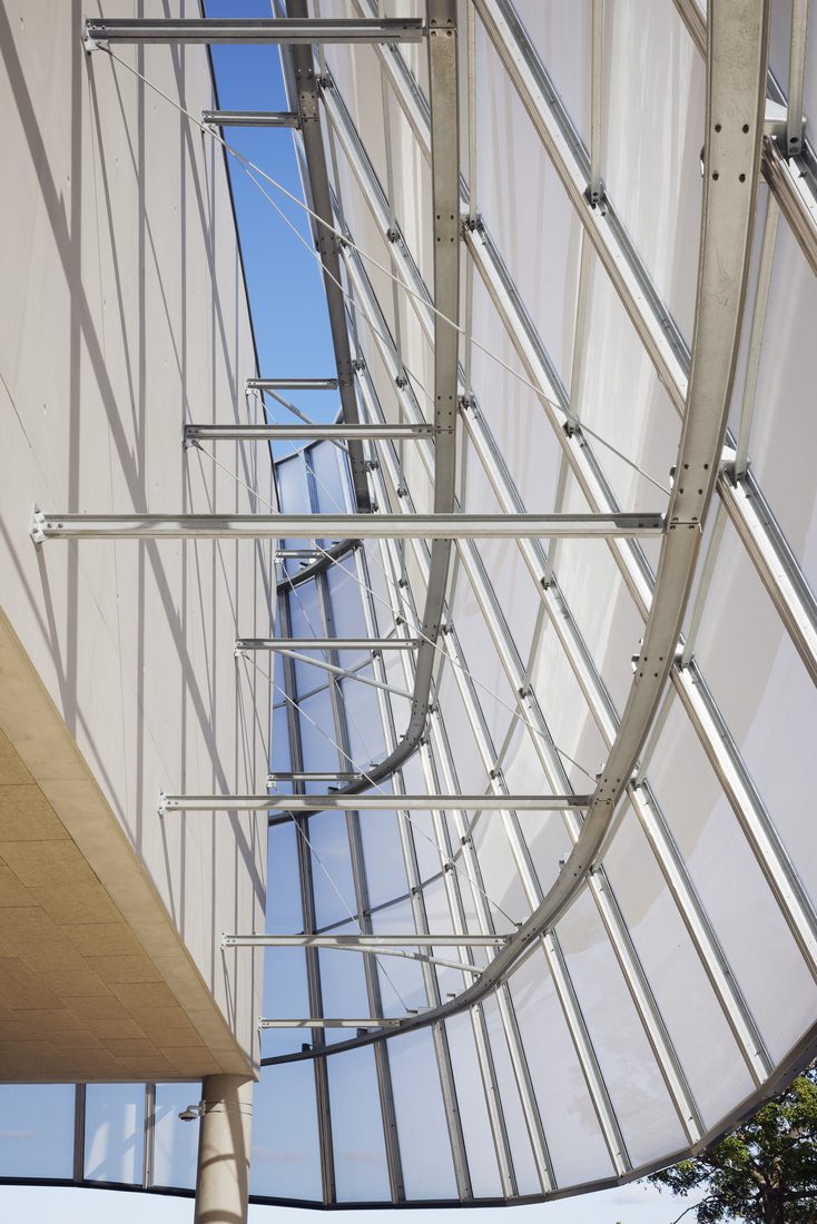 Structure métallique d’une façade courbe en membrane textile translucide, vue en contre-plongée avec ciel bleu en arrière-plan.